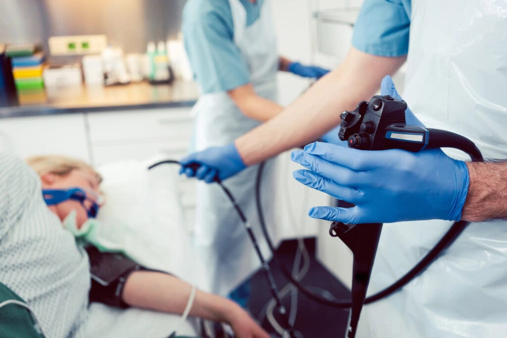 Mediziner führen eine Endoskopie durch. Ein Patient liegt mit einer Sauerstoffmaske auf einem Bett. Ein Mediziner mit blauen Handschuhen hält ein Endoskop, während ein anderer assistiert. Im Hintergrund sind Geräte und Zubehör zu sehen.