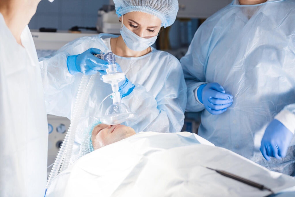 Medizinisches Personal in OP-Kleidung bereitet einen Patienten auf eine Operation vor. Eine Person hält eine Narkosemaske über das Gesicht des Patienten. Andere stehen in der Nähe in einer sterilen Krankenhausumgebung.