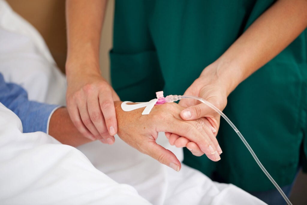 Ein Mitarbeiter in grüner Uniform passt die Infusion in der Hand eines Patienten an. Der Patient liegt mit einem weißen Laken im Bett. Der Mitarbeiter konzentriert sich auf die Platzierung der Infusion und stellt sicher, dass sie sicher sitzt.