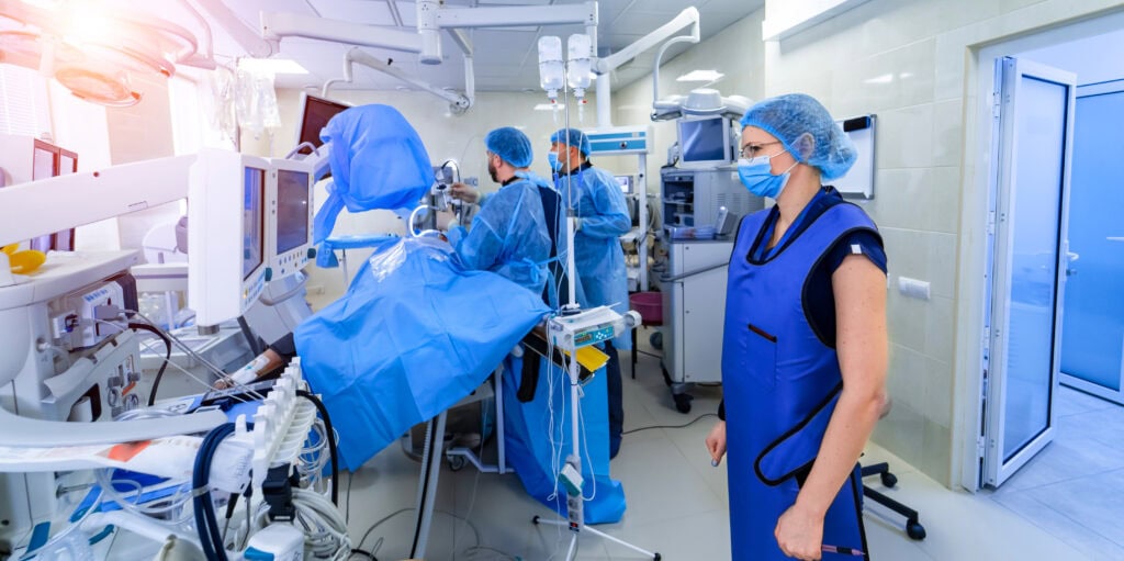 Medizinisches Personal in Schutzkleidung operiert einen Patienten in einem mit hochmodernen Geräten ausgestatteten Krankenhauszimmer. Im Vordergrund steht eine Person mit einer Bleischürze und überwacht den Eingriff.