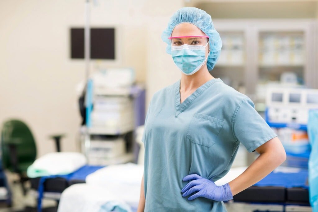 Eine Medizinerin in blauem OP-Kittel, Maske, Handschuhen und Haarkappe steht selbstbewusst mit einer Hand auf der Hüfte in einem mit medizinischem Gerät gefüllten Operationssaal eines Krankenhauses.