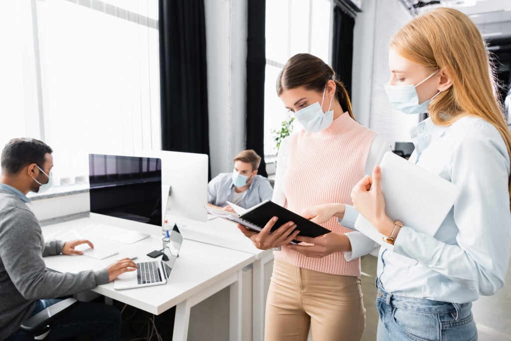 Zwei maskierte Frauen diskutieren, während sie ein Notizbuch in der Hand halten. Eine hat ein Tablet. Im Hintergrund arbeiten zwei maskierte Männer an Schreibtischen mit Computern. Die Kulisse scheint ein modernes Büro mit großen Fenstern zu sein.