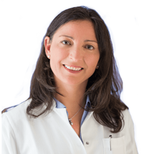 Eine lächelnde Frau mit langem dunklem Haar, einem weißen Arztkittel und einer Halskette bekleidet, steht vor einem schlichten Hintergrund.