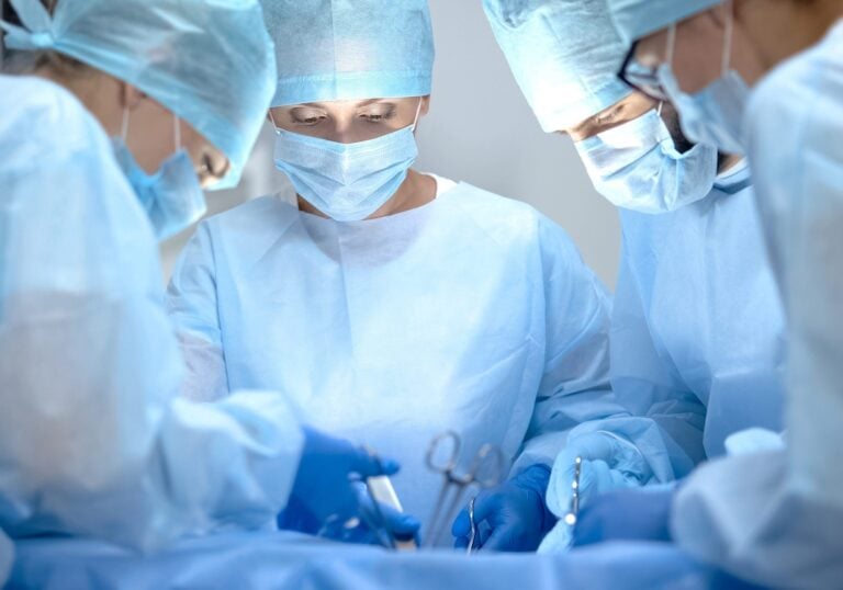 Eine Gruppe von Chirurgen in blauen OP-Kitteln, Masken und Hauben konzentriert sich auf einen Eingriff. Sie stehen eng um einen Patienten versammelt und halten chirurgische Instrumente in der Hand. Der Raum ist hell erleuchtet, was ihre Konzentration und Teamarbeit unterstreicht.