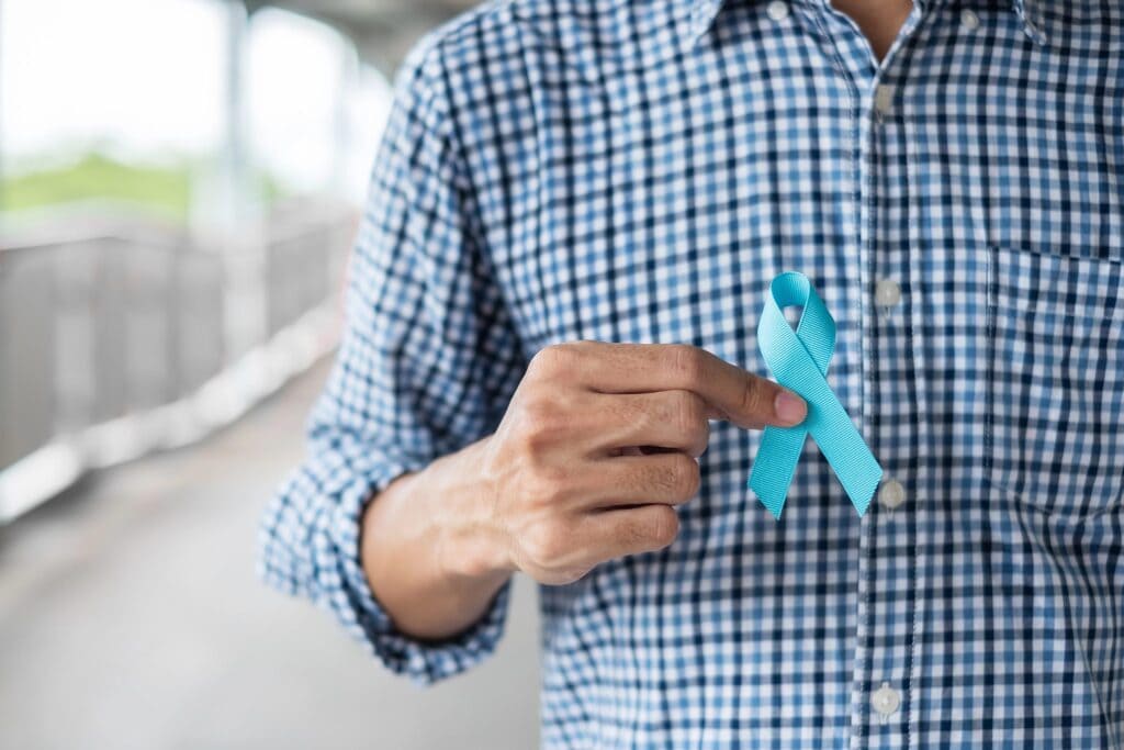 Eine Person in einem blaukarierten Hemd hält ein blaues Band zwischen Daumen und Zeigefinger. Der Hintergrund ist unscharf und lässt auf eine Szene im Freien schließen. Das blaue Band wird typischerweise mit der Aufklärung über Prostatakrebs und Initiativen für Männergesundheit in Verbindung gebracht.