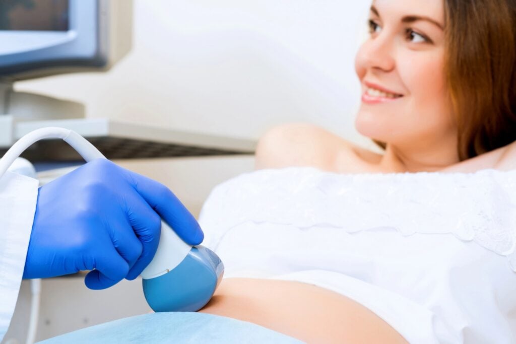 Eine lächelnde Frau liegt auf einem Untersuchungstisch, während eine medizinische Fachkraft mit blauen Handschuhen eine Ultraschalluntersuchung an ihrem entblößten Bauch durchführt.