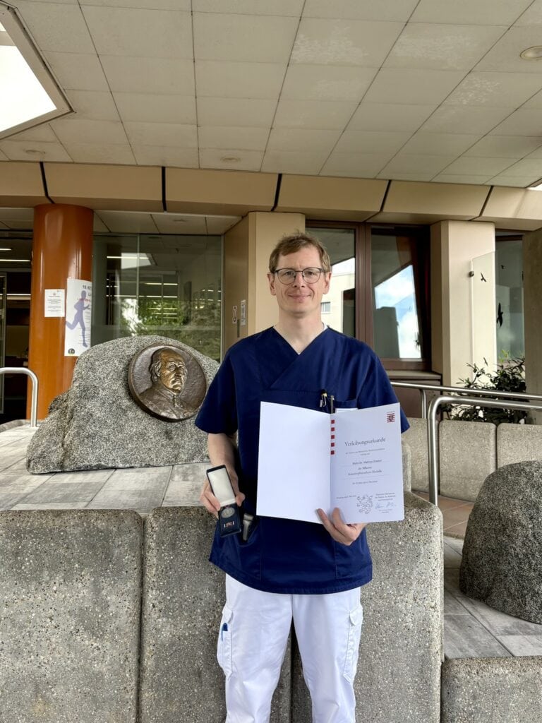Eine Person in blauem OP-Kittel steht vor einem Gebäude und hält ein aufgeschlagenes Zertifikat sowie eine kleine Schachtel mit einer Medaille in der Hand. Hinter ihr befindet sich eine Steinskulptur, an der ein Porträt als Metallrelief angebracht ist.