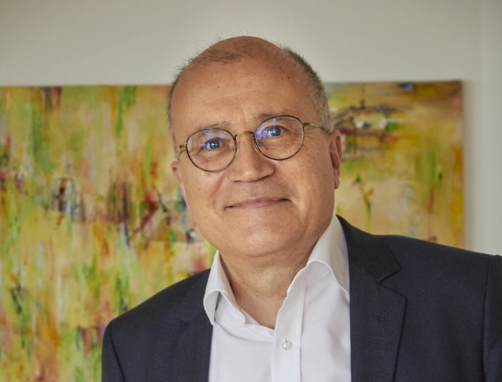 Ein Mann mit Brille und Anzugjacke lächelt vor einem farbenfrohen abstrakten Gemälde an einer weißen Wand.