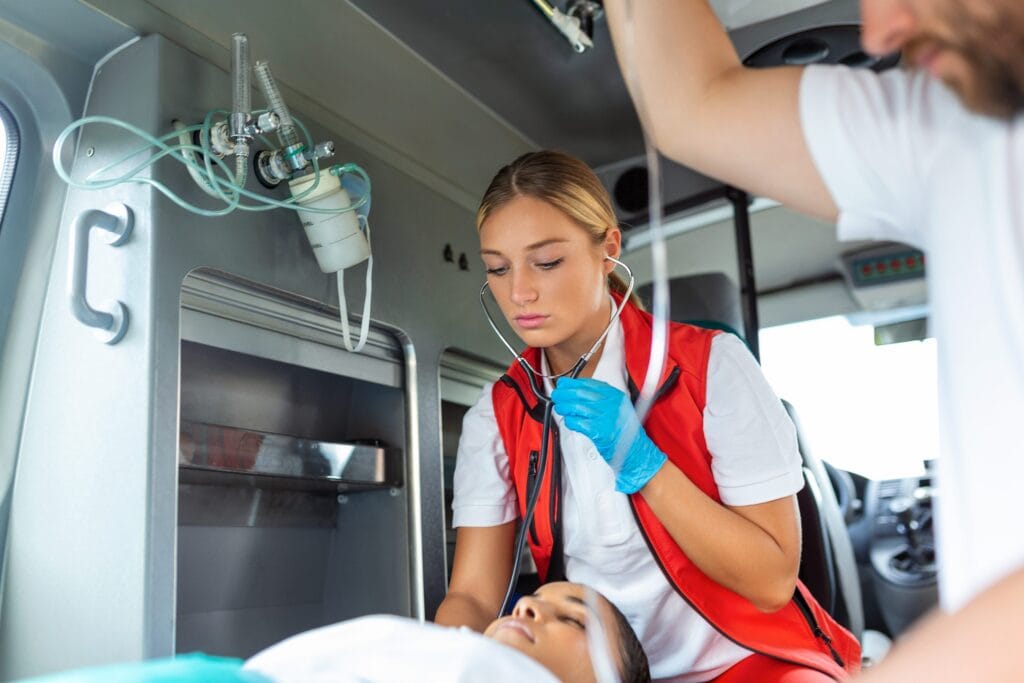 Ein Rettungssanitäter in einer roten Weste untersucht mit einem Stethoskop einen Patienten, der auf einer Trage in einem Rettungswagen liegt, während ein weiterer Rettungshelfer daneben steht und sich an einem Handlauf festhält.